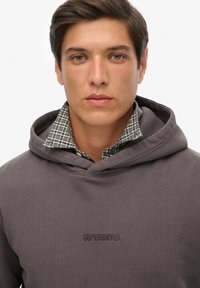 Sudadera marrón con tejido texturizado, que presenta un logo en el pecho. Un collar de camisa de cuadros es visible debajo de la capucha.