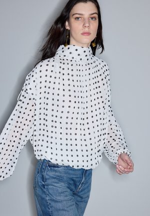 Mujer joven con cabello largo y oscuro, vestida con una blusa blanca de cuello alto con lunares negros y jeans azules, frente a un fondo liso.