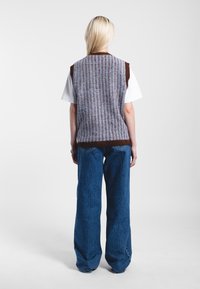 Gilet tricoté rayé gris et marron à manches courtes, porté sur une chemise blanche, associé à un jean en denim bleu à jambe large.