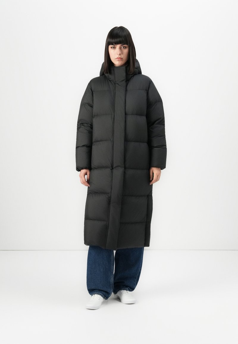 Donna in piedi con un lungo cappotto piumino nero, jeans larghi blu e sneaker bianche su uno sfondo bianco semplice.