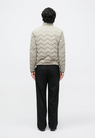 Bézs színű, cikk-cakk mintás, steppelt dzseki, deréknál alakra szabva, fekete nadrággal párosítva. A kialakítás magas gallérral és sima textúrával rendelkezik.