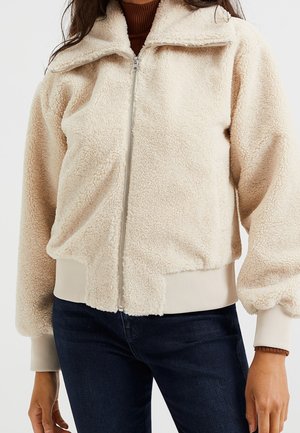Frau trägt eine cremefarbene Fleecejacke mit Reißverschluss und gerippten Bündchen und Saum, kombiniert mit dunkelblauen Jeans.