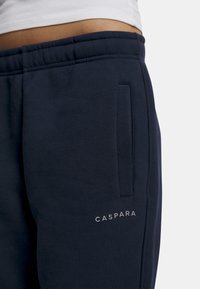 Mørkeblå joggebukser med myk stoff, med en sidelomme og en grå "CASPARA"-logo på underbenet. Elastisk liv. 