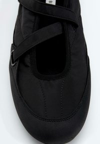 Chaussure plate noire avec bout arrondi, dessus en tissu et bride ajustable traversant le cou-de-pied avec une boucle métallique sur le côté.