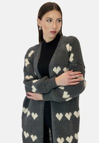Cardigan grigio lavorato a maglia con motivi a cuore bianchi, caratterizzato da un design ampio, maniche lunghe e polsini a coste. Senza chiusure o bottoni.