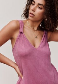 Kvinde med krøllet hår iført guldkæder og en ærmeløs pink top, poserer med den ene hånd på hoften og ser sidelæns.