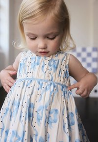 Biele šaty s modrým kvetinovým vzorom a čiernymi bodkami; majú plisovaný dizajn a tenký modrý opasok; dieťa ukazuje na detail opasku.