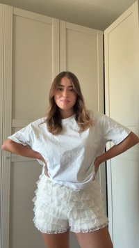 Biała, oversize t-shirt w połączeniu z białymi, falbaniastymi szortami. Posiada koronkowe wykończenie oraz wielowarstwowe tekstury. Neutralne tło.