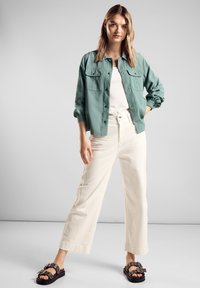 Světle zelená košilová bunda přes bílou košili, doplněná krémovými kalhotami s širokými nohavicemi a tmavými sandály s dekorativními prvky.