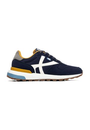 Zapatilla azul marino con logo blanco en el lateral, forro interior amarillo mostaza, talón gris, detalles azules y marrones en la suela y cordones negros.