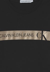 Svart bomullströja med en glansig guldslogo-rand som har "CALVIN KLEIN JEANS" och "CK" tryckt i svart. Crew neck-design.