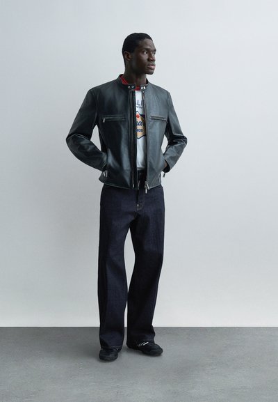 Άνδρας στέκεται με τα χέρια στις τσέπες φορώντας μαύρο δερμάτινο μπουφάν, λευκό μπλουζάκι με σχέδιο, σκούρο χαλαρό τζιν και μαύρα παπούτσια μπροστά σε απλό γκρι τοίχο.