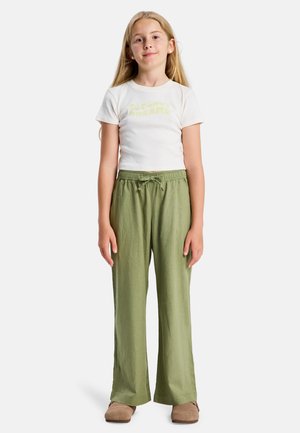 Jong meisje met lang blond haar, een wit shirt met korte mouwen, olijfgroene broek met trekkoord en bruine schoenen, staand met de armen langs het lichaam.