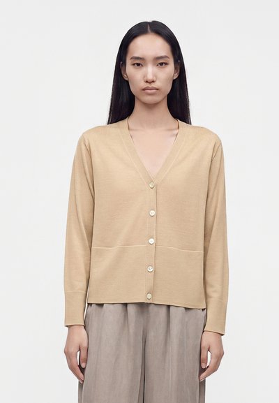 Femme aux longs cheveux noirs portant un cardigan beige boutonné et un pantalon gris ample, debout devant un fond clair uni.