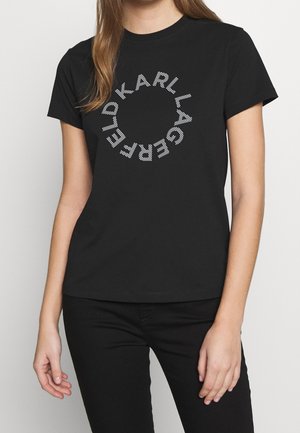 Femme portant un t-shirt noir à manches courtes avec "KARL LAGERFELD" en lettres blanches rayées disposées en cercle sur la poitrine.