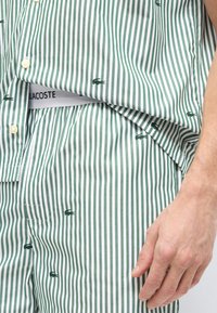 Πράσινο και λευκό πουκάμισο με κάθετες ρίγες και σορτς με μικρά σκοτεινά πράσινα λογότυπα κροκόδειλου, σηκώνοντας μερικώς το πουκάμισο για να αποκαλύψει τη μέση με το κείμενο "LACOSTE".