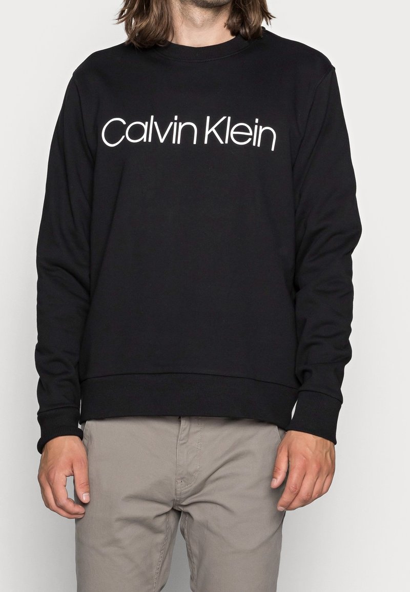 Černá mikina vyrobená z měkké tkaniny, s bílým logem "Calvin Klein" přes hrudník, s žebrovanými manžetami a lemem.