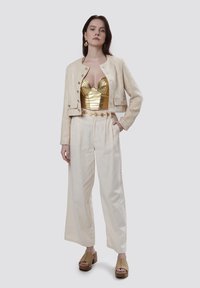 Chaqueta recortada de textura crema, corsé dorado esculpido, pantalones anchos color marfil y sandalias de plataforma marrón. Unos pendientes sencillos completan el conjunto.