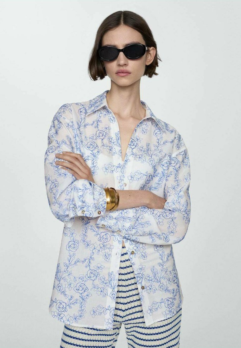 Camicia bianca trasparente con motivo floreale blu, bottone sul davanti e maniche piegate. Il modello indossa occhiali da sole neri e pantaloni a righe.