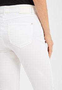 Weiße Jeans mit einem figurbetonten Schnitt, Gesäßtaschen mit silberner Hardware und einem schlichten Taillenlabel. Glatte Textur und keine sichtbaren Muster.