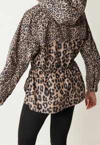 Femme portant une veste à capuche imprimée léopard avec taille froncée et leggings noirs, vue de dos sur un fond uni.