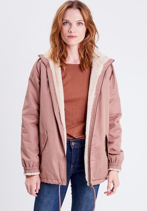 Veste extérieure rose avec un intérieur crémeux et flou, dotée d'une capuche, d'une fermeture éclair à l'avant et de poches latérales. Le modèle porte un top marron et un jean.