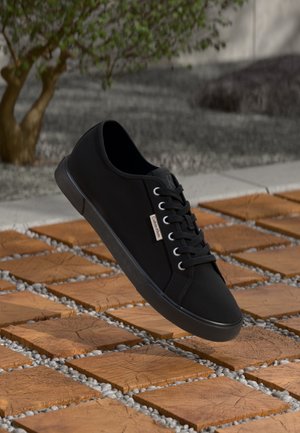 Baskets noires à lacets avec une étiquette Calvin Klein flottant au-dessus de carreaux en bois carrés en extérieur, avec un arbre et du gravier en arrière-plan.