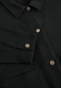 Camicia nera in tessuto testurizzato con colletto, bottoni a pressione dorati e tasca, che mette in evidenza un design a nido d'ape.