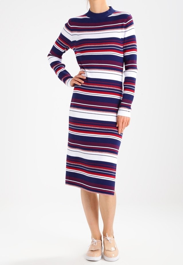 Femme portant une robe ajustée à manches longues, longueur genou, avec des rayures horizontales bleu marine, blanc, rouge et bordeaux, et des chaussures beiges à lacets.