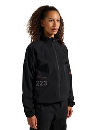 Woman with curly hair wearing a black zip-up jacket with printed numbers and letters, standing against a white background.