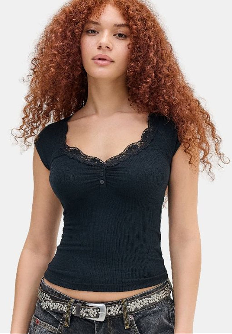Junge Frau mit langen, lockigen roten Haaren, die ein eng anliegendes schwarzes Top mit Spitzenbesatz und einen mit Nieten besetzten schwarzen Gürtel über dunklen Jeans trägt.