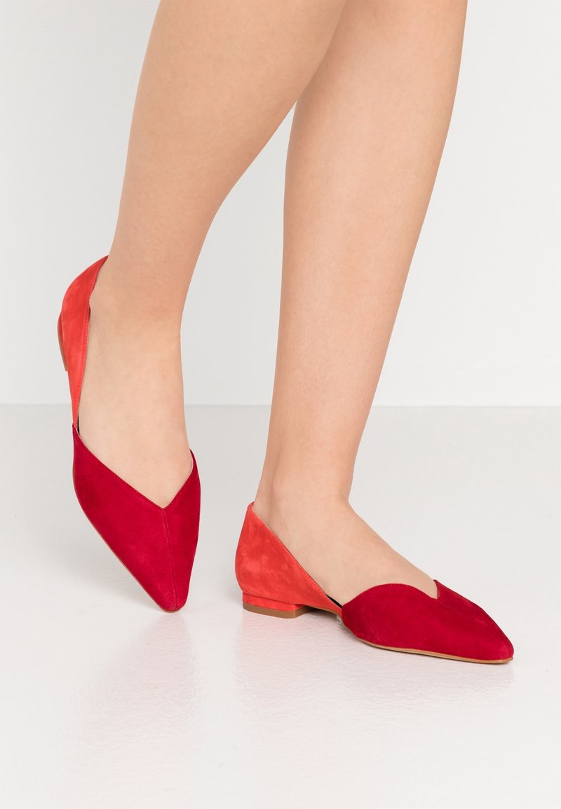Jambes portant des ballerines en daim bicolores rouge et corail à bouts pointus avec petits talons, sur un fond de sol et de mur clair et uni.