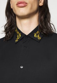 Camisa negra con un frente negro abotonado, dos botones y un patrón amarillento bordado en el cuello. Textura de tela suave.