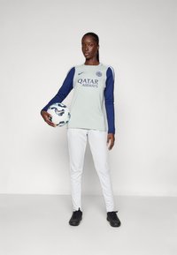 Un maillot à manches longues gris clair avec des bras navy et le texte "QATAR AIRWAYS", associé à un pantalon blanc et des chaussures de sport noires, tenant un ballon de football à motifs.