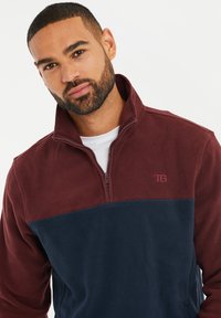 Pullover in fleece con colletto a zip, caratterizzato da una parte superiore bordeaux e una parte inferiore blu navy. Include un piccolo logo sul petto.