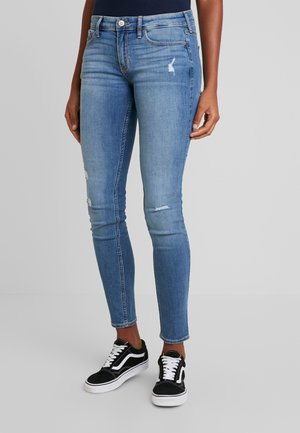 Kobieta w jasnoniebieskich obcisłych jeansach z lekkim przetarciem, w połączeniu z czarno-białymi sneakersami na sznurowadła, na jednolitym szarym tle.