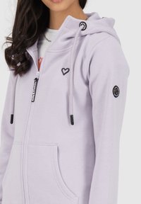 Felpa con zip di colore viola chiaro, con logo a forma di cuore sul petto, cappuccio con lacci e dettagli neri. Presenta tasche frontali e un tessuto testurizzato.