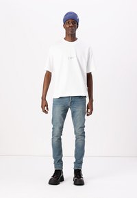 White short-sleeve t-shirt with small front logo, paired with light blue skinny jeans and black lace-up boots, complemented by a blue beanie.