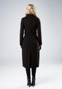 Blondynka odwrócona tyłem, ubrana w długi czarny płaszcz z paskiem, czarne spodnie i czarne botki na obcasie, na jednolitym tle.