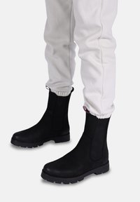 Sorte elastiske ankelsko med en glat overflade, rund tå, lav blokhæll, og side træksløjfer, parret med lysfarvede sweatpants.