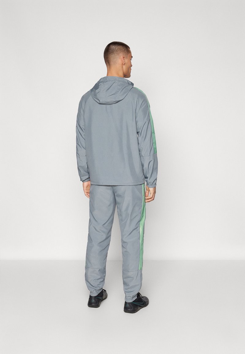 Ensemble de sport gris composé d'une veste à capuche et d'un pantalon assorti, avec des rayures latérales vertes. Tissu lisse et léger.