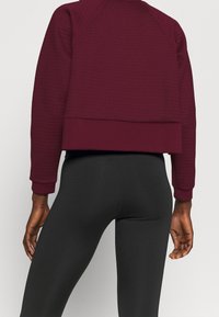 Persona che indossa un maglione a maniche lunghe texture color bordeaux e leggings neri aderenti, mostrata di spalle con le mani rilassate ai lati.