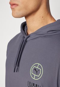 Grå hettegenser med snor, med et sirkulært logo i neongrønn og svart. Glatt stoff med en avslappet passform.