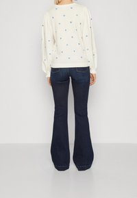 Vrouw in een witte trui met lange mouwen, versierd met kleine blauwe bloemen, en een paar donkere spijkerbroeken met wijde pijpen, staand met haar rug naar de camera tegen een effen achtergrond.