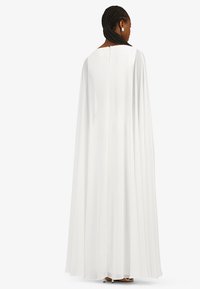 White floor-length gown featuring a soft, flowing cape extending from the shoulders, with a discreet back closure and smooth texture.