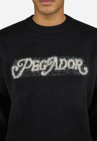 Black sweater with a fuzzy white "PEGADOR" text logo. The knit texture is soft, featuring a round neckline and ribbed collar.