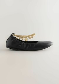Chaussure ballerine noire avec côtés élastiques et bride de cheville en chaîne dorée ornée de breloques en forme d'étoile et de nœud sur fond uni.