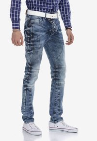 Vervaagde blauwe denim jeans met contrasterende stiksels, strak ontwerp, meerdere zakken, en een witte riem gedragen met witte sneakers.