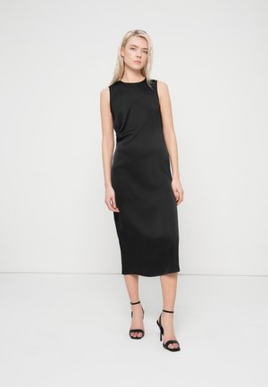 Robe midi noire sans manches, tissu lisse, col rond, design légèrement drapé, portée avec des talons à lanières noirs. Silhouette simple et ajustée.