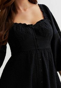 Svart texturerad klänning med knappstängning, fyrkantig halsringning och långa ärmar som har intrikata vävda mönster. Mjuk tygkvalitet med en avslappnad silhuett.